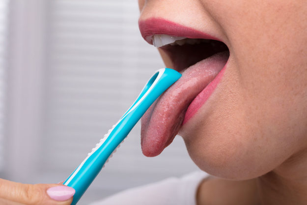 causes of bad breath (4) woman using tongue scraper to prevent causes of bad breath